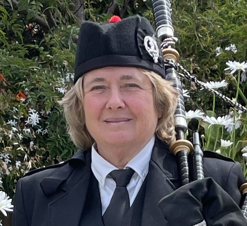 Person in traditional Scottish attire holding bagpipes, surrounded by flowers.