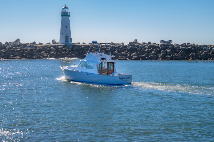 White boat on water near a lighthouse and rocky shore.