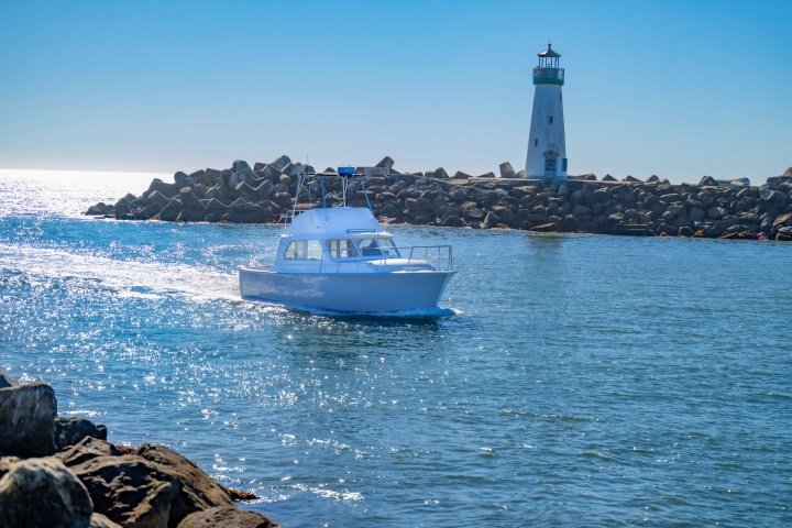 White boat near lighthouse on rocky shore under clear blue sky.